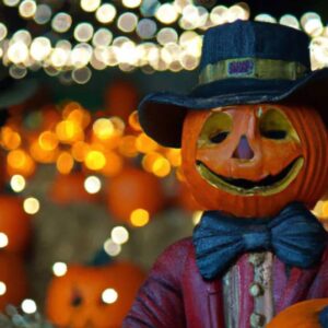 A jack 'o lantern pumpkin head with a hat stands in front of festival lights
