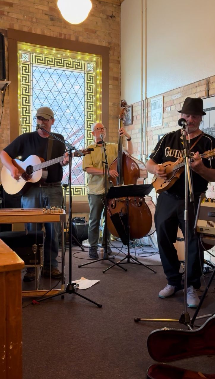 The Dawg performing on a small stage, playing guitar, bass, and mandolin in front of a decorative glass window and brick wall.