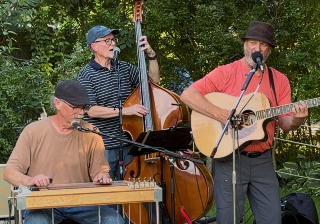A Dawg concert performing outdoors in front of lush green trees on, a steel guitar, bass, and guitar while singing their signature tight harmonies.